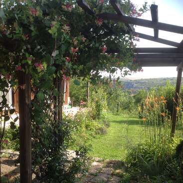 The image depicts a serene garden scene with a wooden pergola, lush greenery, and vibrant flowers, set against a picturesque backdrop of rolling hills.