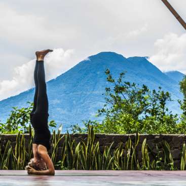 The image depicts a woman performing a headstand on a yoga mat, set against a backdrop of a mountain range and lush greenery.
