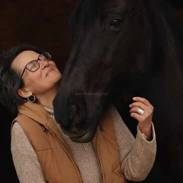 The image depicts a woman with short dark hair, wearing glasses and a tan vest, tenderly embracing a black horse\'s face. The woman\'s hand is gently touching the horse\'s nose.