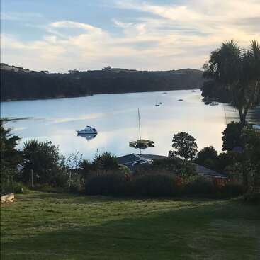 La imagen representa una serena escena lacustre con barcas, rodeada de frondosa vegetación y árboles, bajo un cielo nublado. En primer plano se ve una casa.