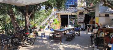 The image depicts a serene outdoor patio area, featuring a table and chairs, surrounded by lush greenery and a staircase leading to a building with a balcony.