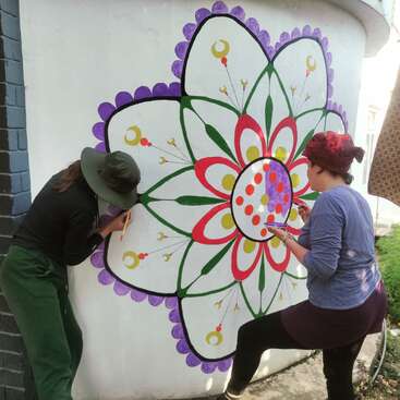 Two women are painting a large flower mural on a white wall, using paintbrushes and vibrant colors, with a brick wall and grass visible in the background.
