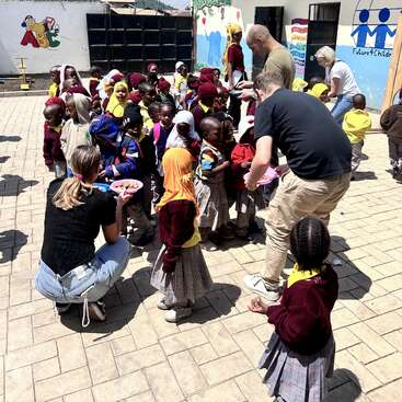 A group of young children in uniforms gathers outside, interacting joyfully with adults distributing food or treats near a colorful, mural-decorated building under a bright sky.