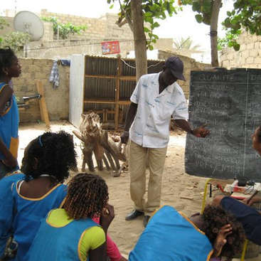 The image depicts a man teaching a group of children outside, using a chalkboard to illustrate a lesson, with a serene background of trees and a stone wall.