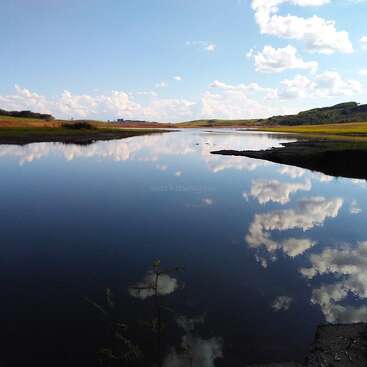 The image depicts a serene landscape featuring a body of water, reflecting the sky and clouds, with lush greenery on either side and a blue sky above.