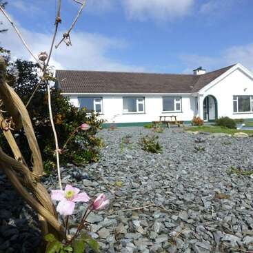 The image depicts a white house with a brown roof, surrounded by a gravel yard and a wooden bench, set against a blue sky with white clouds.