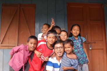 Das Bild zeigt einen Mann, der mit einer Gruppe von Kindern vor einem Gebäude posiert. Der Mann trägt eine Brille und ein gestreiftes Hemd, während die Kinder Freizeitkleidung tragen.