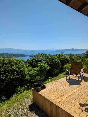 The image depicts a serene wooden deck with a bucket and chair, overlooking a tranquil body of water, surrounded by lush greenery and majestic mountains in the distance.