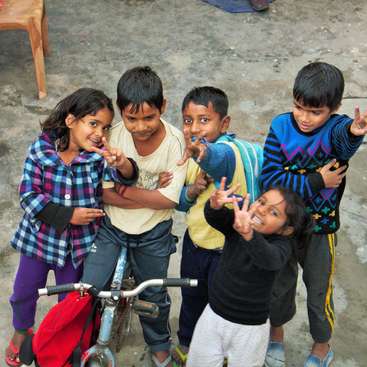 The image shows five children standing around a bicycle, posing for the camera with their hands in various peace signs. They are all wearing casual clothing.