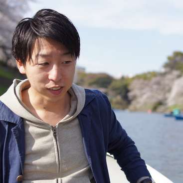 The image depicts a young man with short black hair, wearing a blue jacket over a gray hoodie, standing in front of a serene body of water and a blurred landscape.