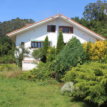 The image depicts a white house with a red roof, surrounded by lush greenery and trees, set against a clear blue sky on a sunny day.