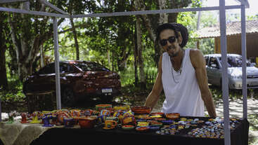 The image depicts a man standing behind a table, with a bookshelf in the background. The man is wearing a white shirt and has short hair. The scene appears to be a domestic setting.