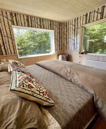 The image depicts a cozy bedroom with two beds, adorned with brown bedding and patterned pillows, featuring large windows that offer a serene view of trees outside.