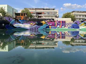 The image depicts a vibrant mural of a dragon on a wall, reflected in water, with a building and trees in the background, set against a blue sky with clouds.