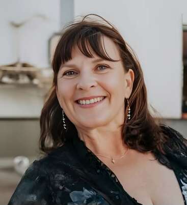 The image depicts a woman with fair skin and brown hair, wearing a black top and dangling earrings, smiling at the camera with a blurred background.