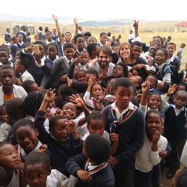 The image depicts a joyful scene of a large group of children and two adults posing for a photo in a rural setting, with a vast open field and hills in the background.