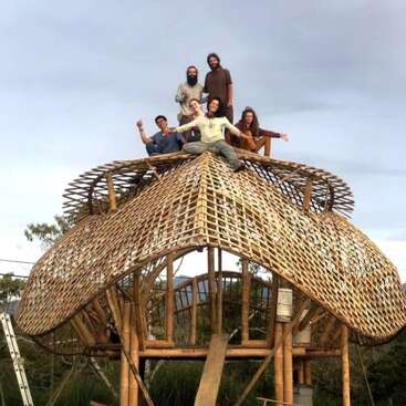 The image depicts a group of people standing on top of a large bamboo structure, with one person sitting on the roof, set against a cloudy sky.