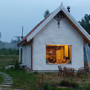 The image depicts a white house with a red roof, featuring a large window that reveals a cozy interior, set amidst a serene natural landscape.