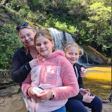 L'image représente une femme et deux jeunes filles posant devant une cascade, la femme se tenant derrière les jeunes filles, qui sont assises sur un rocher.