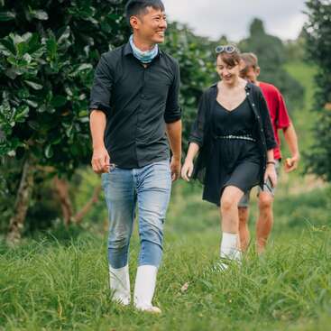 The image depicts three individuals strolling through a lush grassy field, surrounded by trees, with two of them wearing white boots and the third sporting a red shirt.
