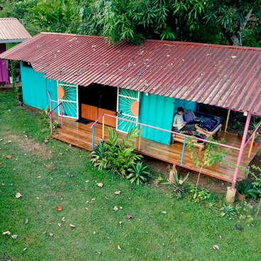 A small, colorful house with a corrugated metal roof sits in lush greenery. A person relaxes on the porch, surrounded by plants and tropical trees.