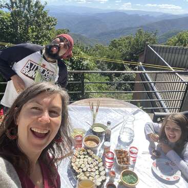 The image depicts a family of three enjoying a meal on a balcony with a scenic mountain view, surrounded by trees and a bright blue sky.