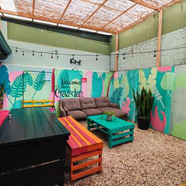 A vibrant outdoor lounge area features colorful pallets, a cozy sofa, potted plants, string lights, and a mural reading “Welcome to Hello’s Garden” on painted walls.