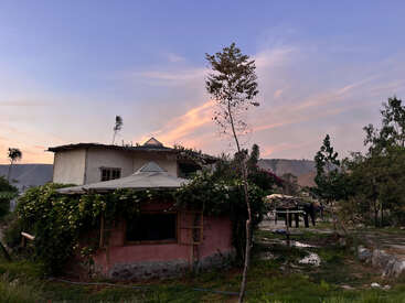 The image depicts a two-story house with a pink exterior, a white roof, and lush greenery, set against a serene mountainous backdrop at sunset.
