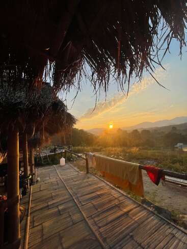 The image depicts a serene sunset scene from a wooden deck, featuring a thatched roof and a railing adorned with towels, set against a picturesque mountainous backdrop.