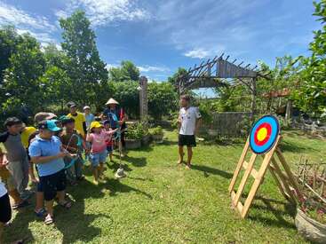 The image depicts a group of children and an adult standing in a grassy area, engaged in archery practice with bows and arrows, near a target on a wooden stand.
