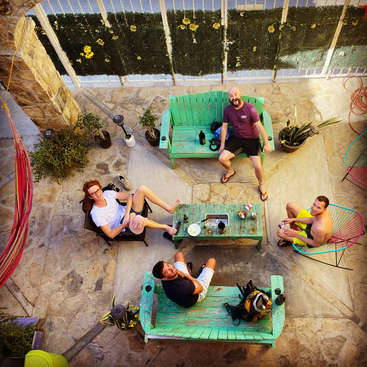 The image depicts four men sitting on a patio, surrounded by furniture and plants, with a stone wall and fence in the background, viewed from above.