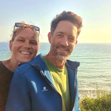 The image shows a man and woman posing for a selfie in front of a body of water, with the man wearing a blue jacket and the woman wearing a black shirt.