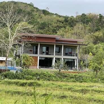 The image depicts a two-story house with a balcony, situated amidst lush greenery and trees, with a serene hillside and overcast sky in the background.