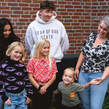 A group of six people, including children and adults, stand together outdoors, smiling and interacting happily in front of a brick wall. Casual and cheerful atmosphere.
