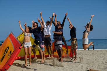 The image depicts a group of eight people jumping on the beach, with their arms raised in the air, in front of a yellow and red inflatable raft.