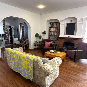 This cozy living room features a patterned sofa, yellow blanket, armchairs, coffee table, bookshelf, fireplace, wooden floor, houseplants, and arched entryways to the kitchen.