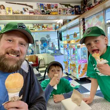 A imagem mostra um homem e dois meninos comendo casquinhas de sorvete em uma sorveteria com brinquedos nas paredes e uma máquina de garra ao fundo.