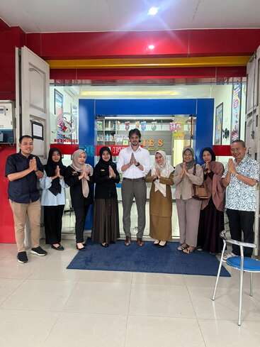The image depicts a group of nine people standing in front of a storefront, with eight individuals wearing headscarves and one man in a white shirt, all with their hands clasped together in a prayer-like gesture.