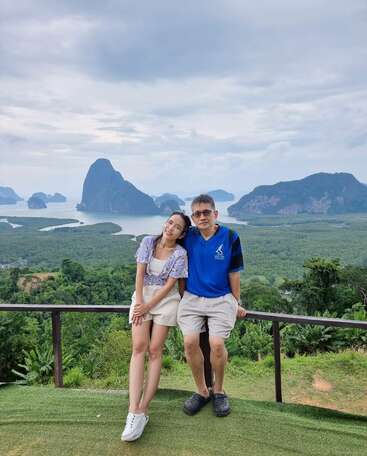 The image depicts a man and woman posing for a photo on a balcony overlooking a lush landscape with distant mountains and water, set against an overcast sky.