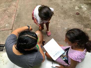 The image shows three young girls sitting on the ground, engaged in an activity with paper and writing utensils, viewed from above.
