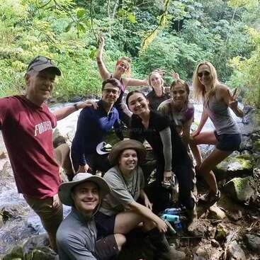 A group of nine people pose happily together in a lush green forest by a stream, smiling, making peace signs, and enjoying an outdoor adventure.