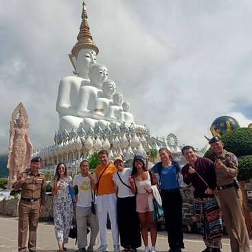 A group of smiling people pose in front of the impressive five white Buddha statues at Wat Pha Sorn Kaew temple, with lush greenery and cloudy sky.