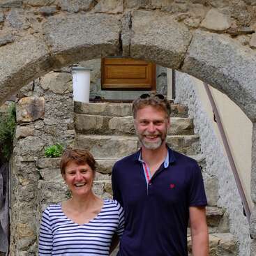 The image depicts a man and woman standing together in front of a stone staircase, with the man wearing a blue polo shirt and the woman in a striped top.
