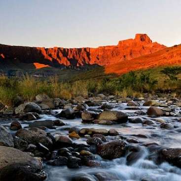 The image depicts a serene landscape featuring a rocky riverbed, lush greenery, and a majestic mountain range in the background, bathed in warm sunlight.