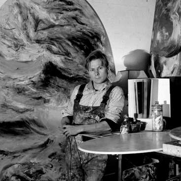 The image depicts a man in overalls standing in front of a large abstract painting, with a table and various art supplies nearby, in a studio setting.