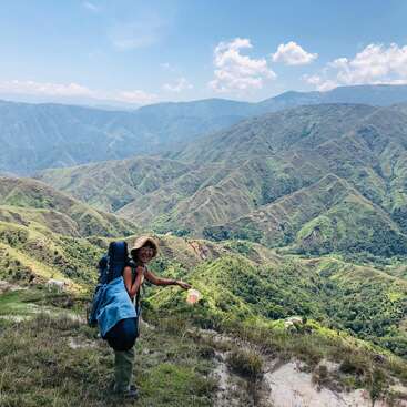 A woman stands on a mountain trail, wearing a backpack and hat, holding a water bottle, with rolling green hills and mountains in the background under a blue sky with white clouds.