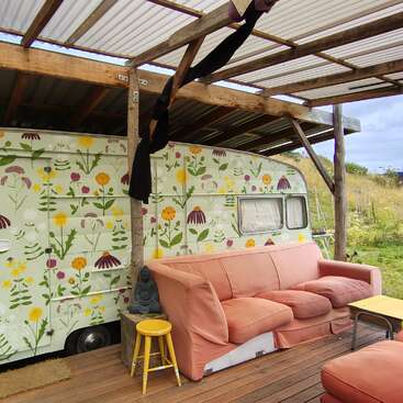 A cozy outdoor space features a covered porch, pink sofa, floral-painted camper, yellow stool, Buddha statue, and green scenery, creating a relaxed and artistic atmosphere.