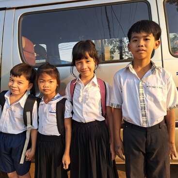 The image shows four children standing in front of a white van, wearing school uniforms and backpacks, with a serene background of trees and power lines.