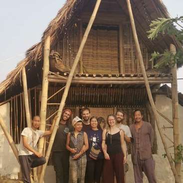 The image depicts a group of eight people standing in front of a bamboo house, with a ladder leaning against the structure and a clear sky in the background.