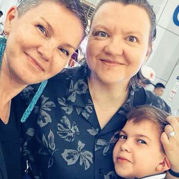 A happy group photo of two women and a young boy, all smiling warmly. The background suggests an indoor public space. Bright, casual, joyful moment.